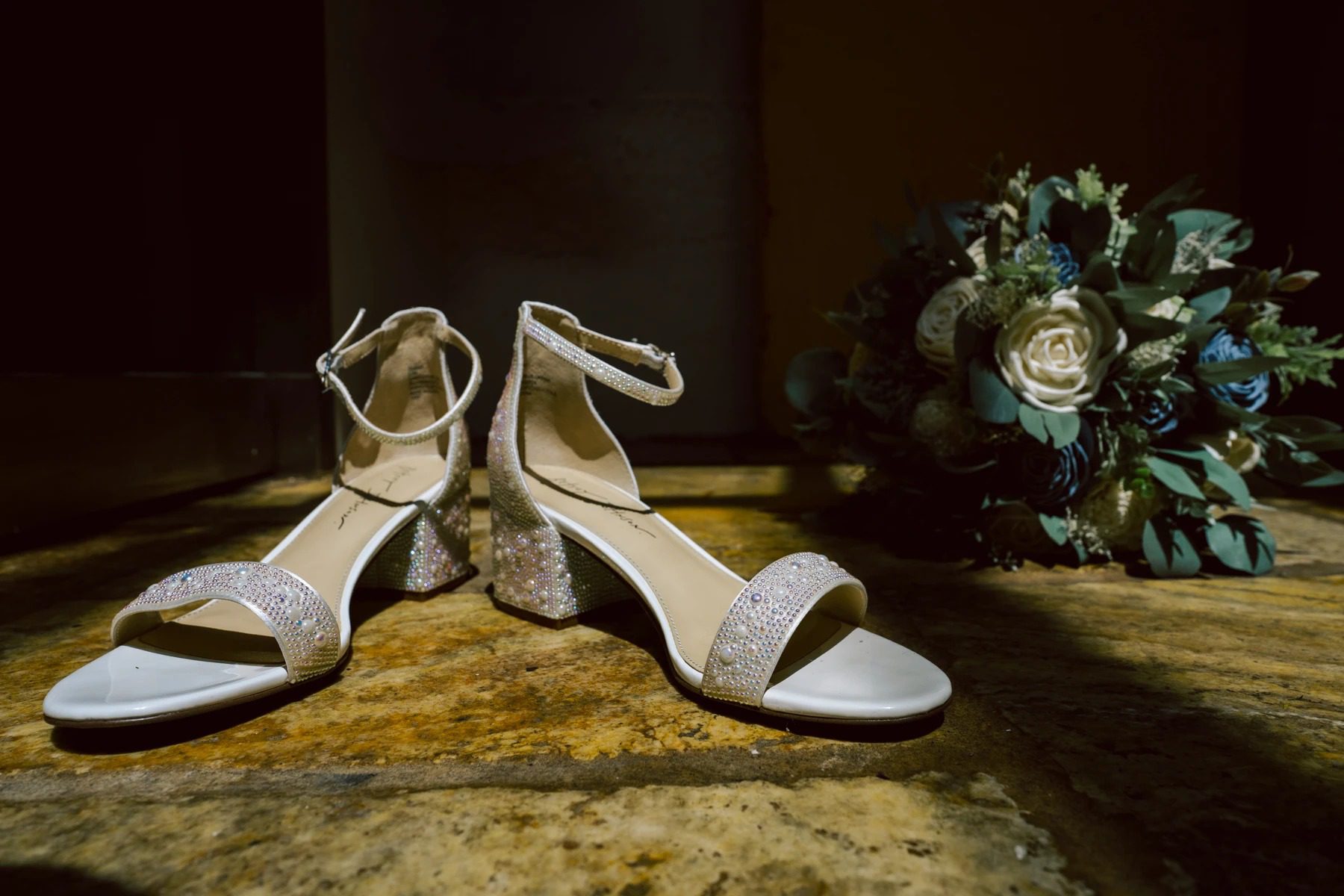 Bride's white embellished flat sandals displayed on stone floor beside bridal bouquet. — Wedding at Dramatic Hall, peekskil, ny