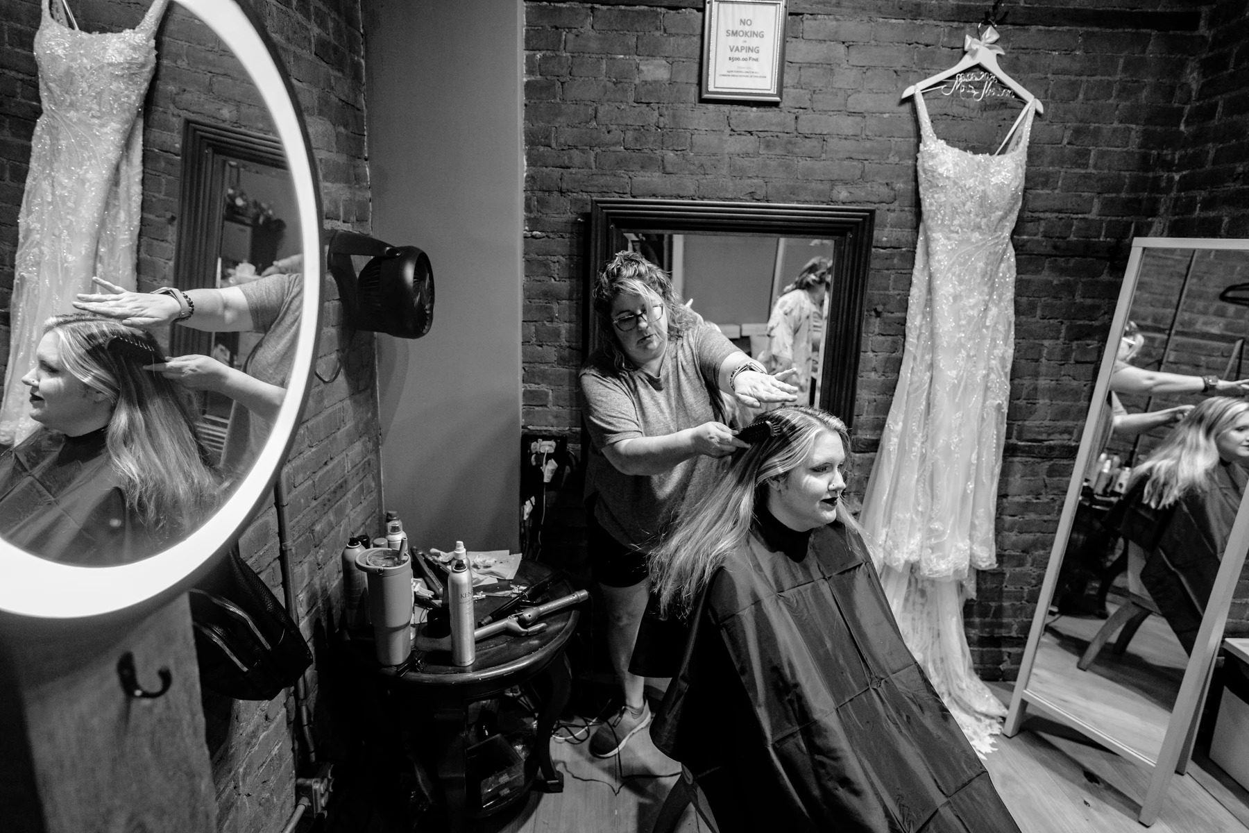Hairstylist curling a woman's hair while she sits in a salon chair, with wedding dresses hanging nearby. — Wedding at Dramatic Hall, peekskil, ny