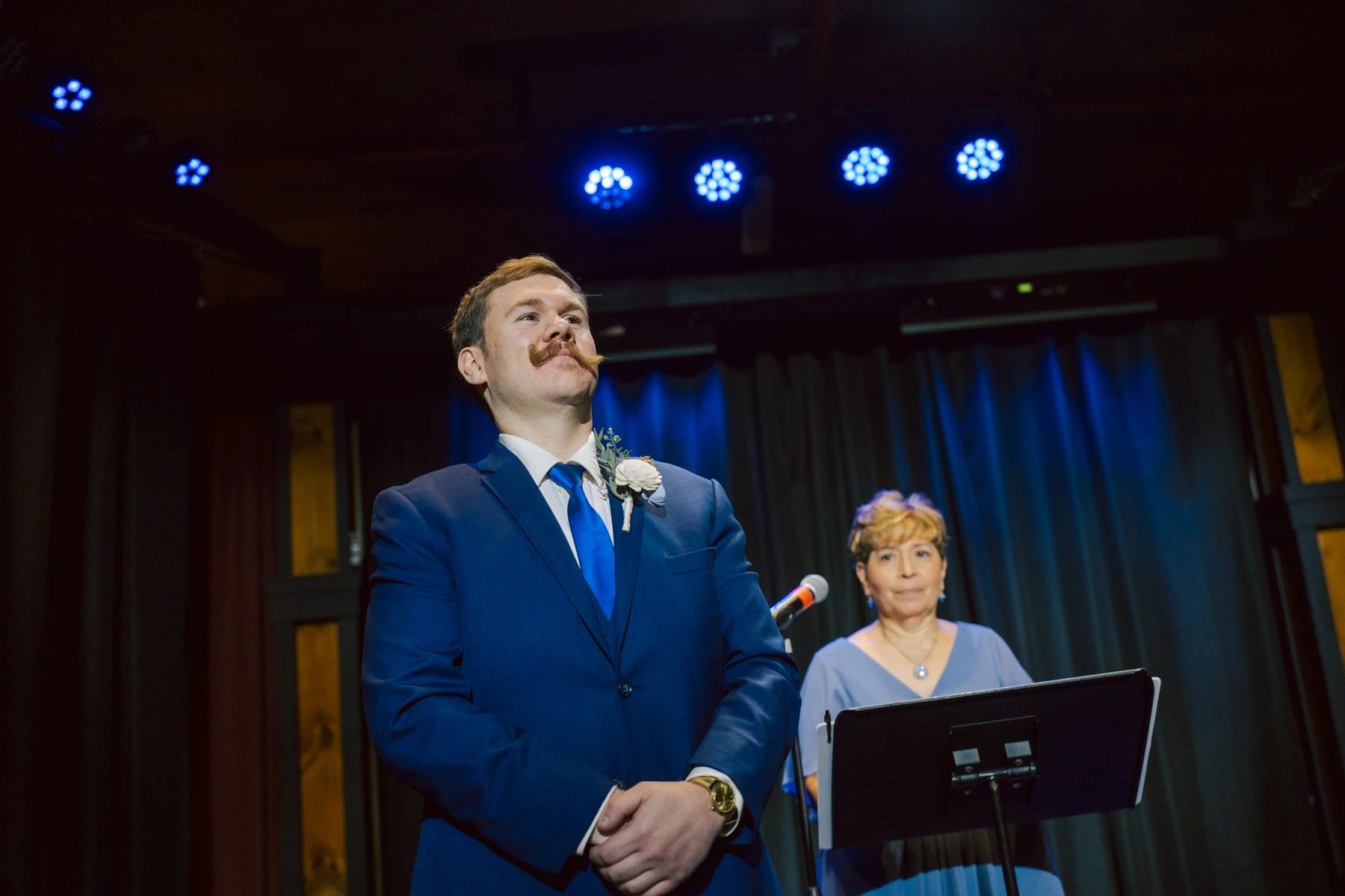 Man in navy suit speaking at microphone while woman stands beside him holding a tablet during reception speech. — Wedding at Dramatic Hall, peekskil, ny